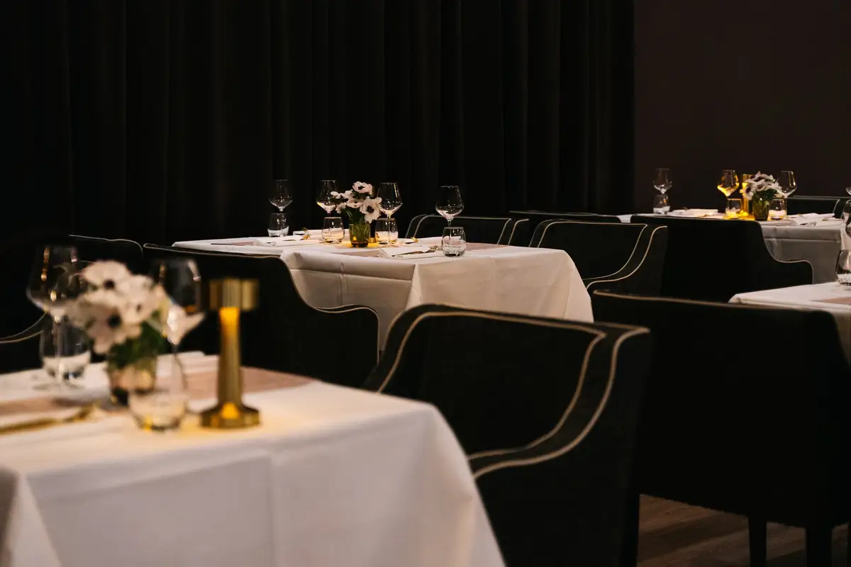 Donker restaurant met gedekte tafels, wit linnen, witte anemonen en een gouden tafellamp voor zwarte gordijnen.