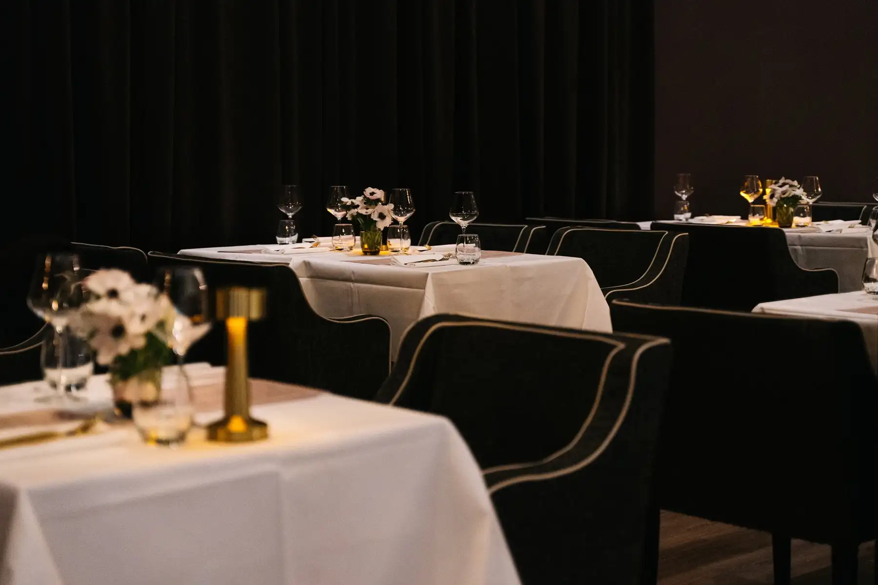 Donker restaurant met gedekte tafels, wit linnen, witte anemonen en een gouden tafellamp voor zwarte gordijnen.