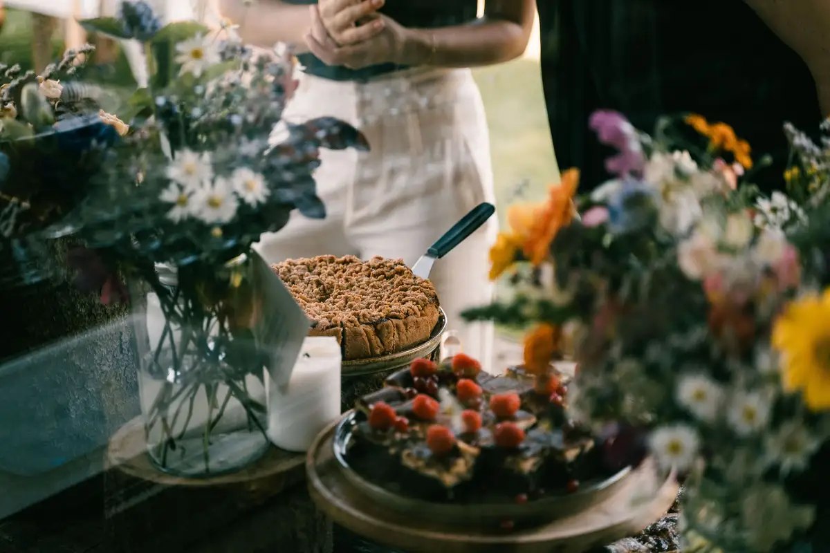 Kruimeltaart en brownies met frambozen tussen veldbloemen in glazen vazen, met een kaars op een houten buffet.