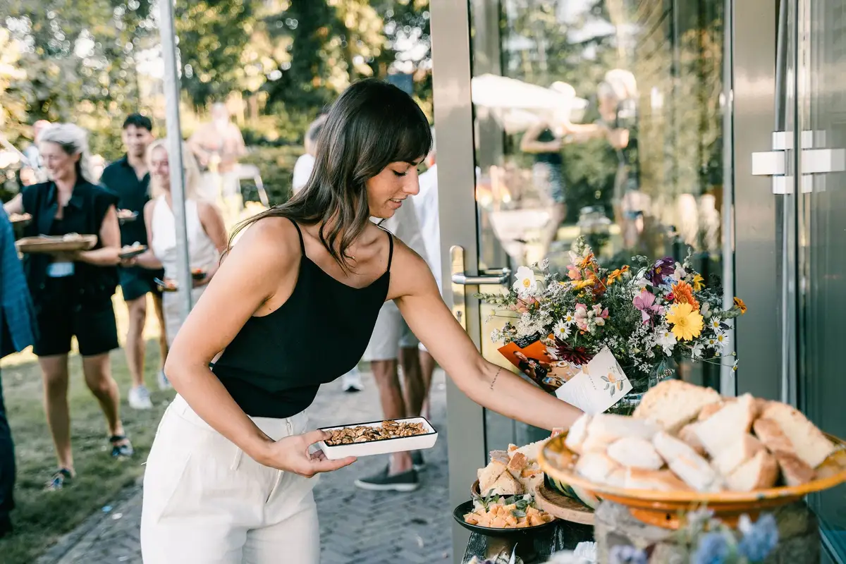 Stella zet een schaal met hapjes bij het buffet in de tuin; gasten lopen erachteraan.