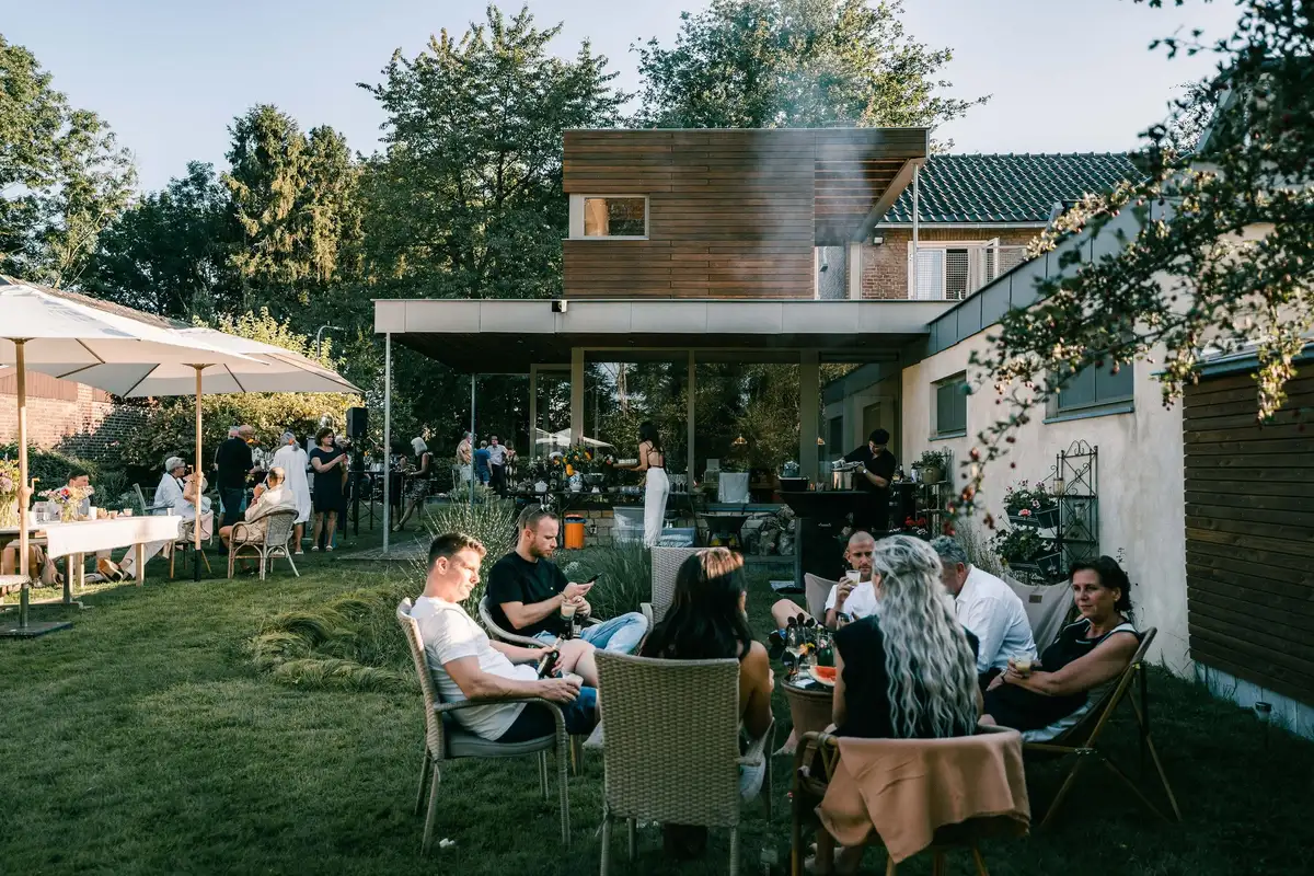 Gasten in de tuin onder parasols en aan lange tafels, achter hen de open keuken van het huis in de avondzon.
