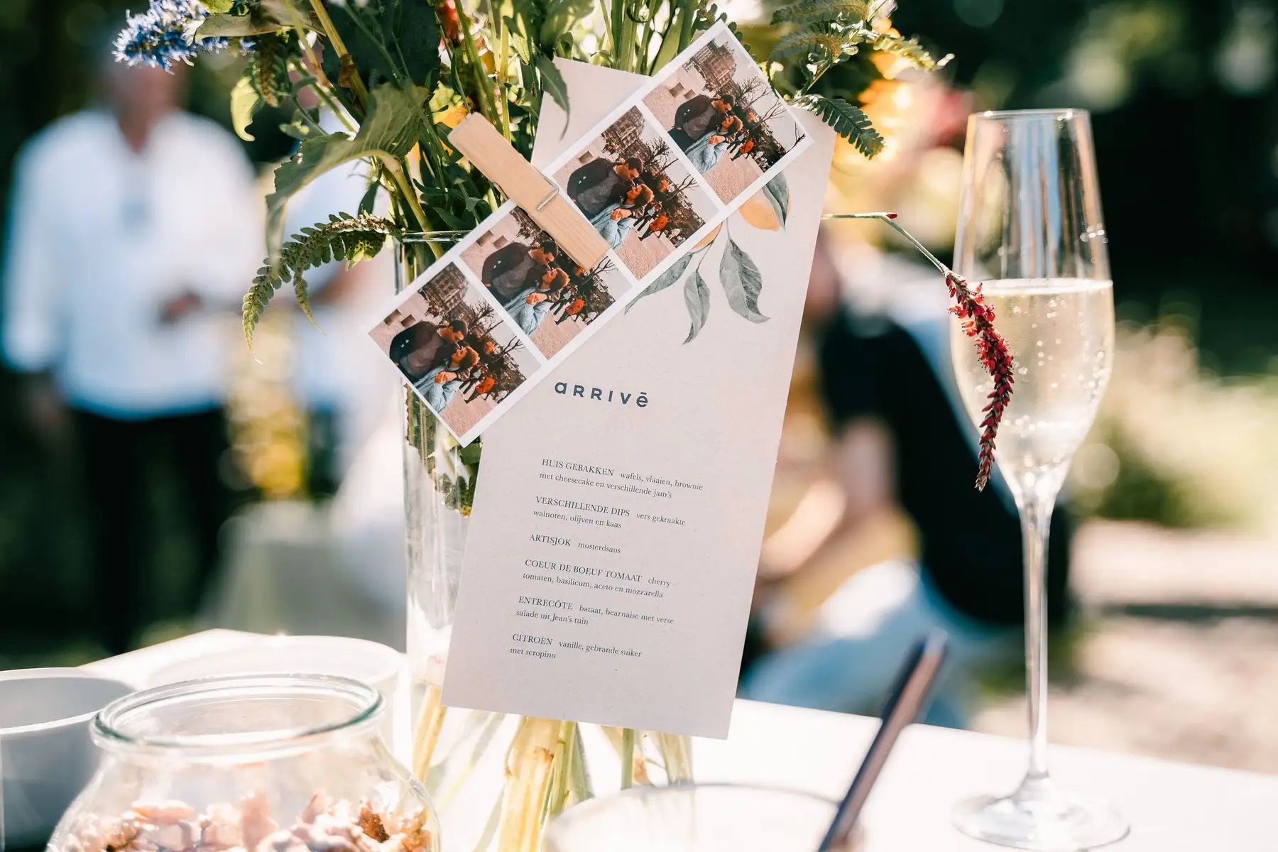Menukaart van Arrivé met fotostrip en citroentak aan een vaas met veldbloemen, naast een glas bruisende wijn in de zon.