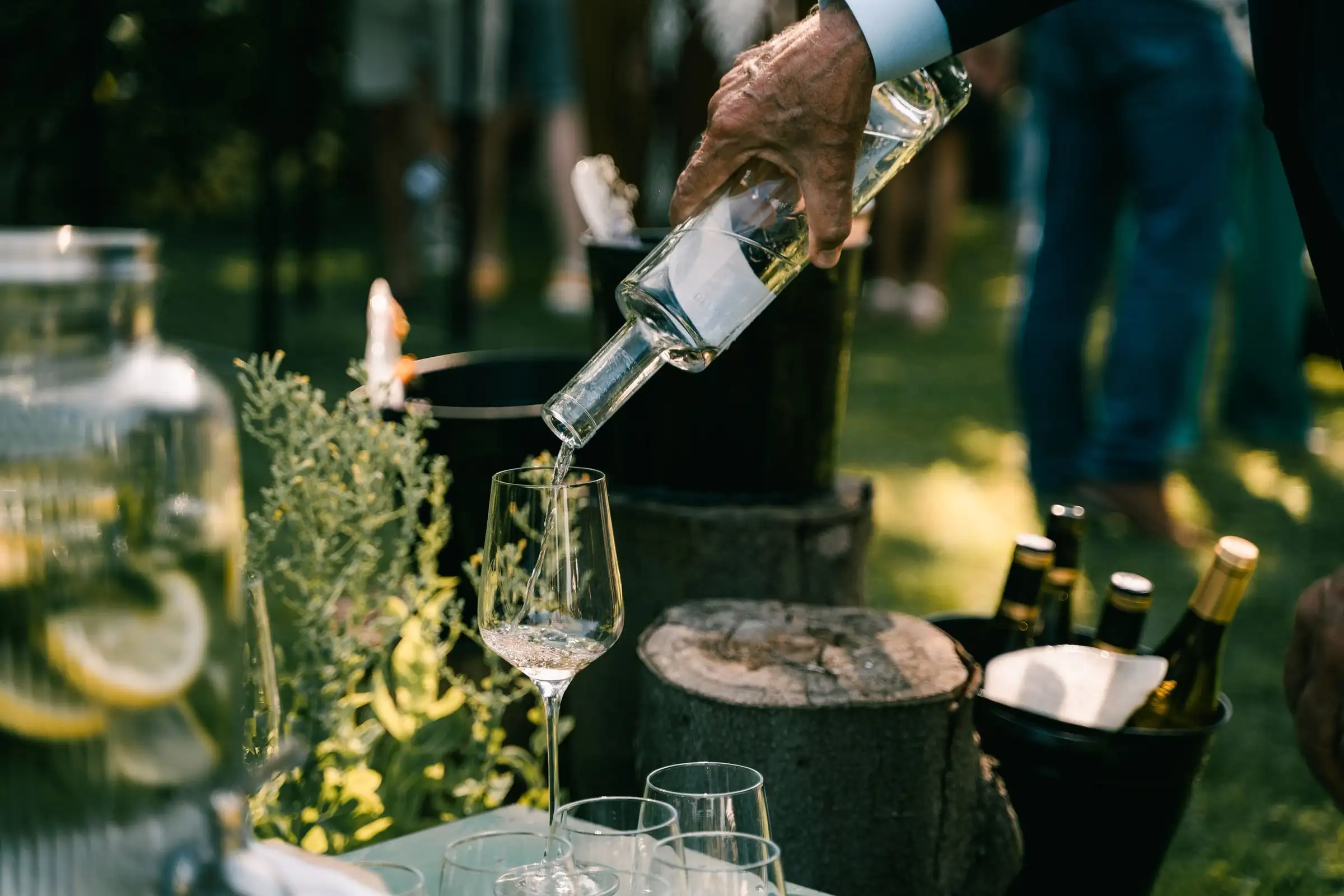 Een hand schenkt witte wijn in een glas op een boomstamtafel in de tuin.