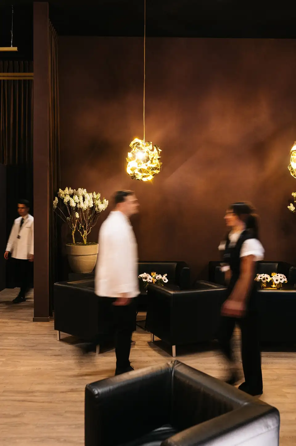 Twee medewerkers lopen in beweging door een warme zaal met een gouden hanglamp.